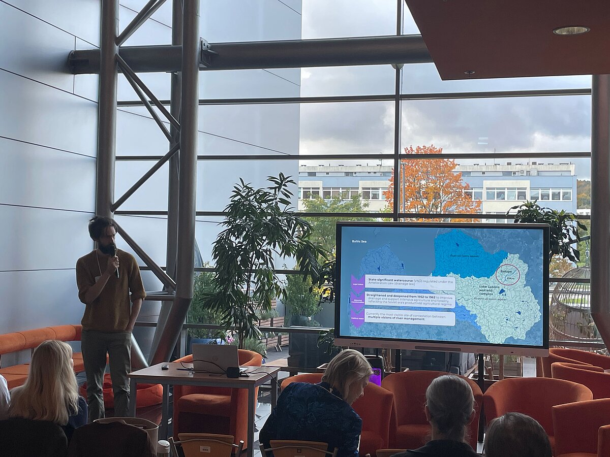 The image on the left shows project researcher Kārlis Lakševics. The image on the right shows a presentation slide. The slide shows a map of Latvia with text annotations. In the background, there is a glass wall, indoor plants, and a view of the neighboring buildings. The audience is visible in the foreground. The image on the left shows project researcher Kārlis Lakševics. The image on the right shows a presentation slide. The slide shows a map of Latvia with text annotations. In the background, there is a glass wall, indoor plants, and a view of the neighboring buildings. The audience is visible in the foreground.