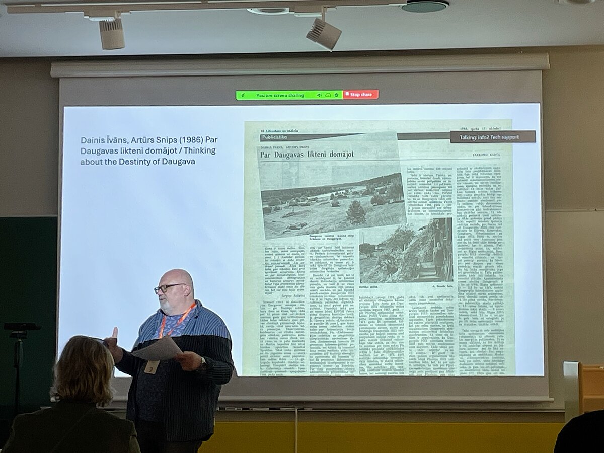 In the picture, a presentation slide with the text "Dainis Īvāns, Artūrs Snips (1986) Thinking about the fate of the Daugava" on the left and a newspaper article on the right, presented by the leading researcher Artis Svece, located on the left side. In the picture, a presentation slide with the text "Dainis Īvāns, Artūrs Snips (1986) Thinking about the fate of the Daugava" on the left and a newspaper article on the right, presented by the leading researcher Artis Svece, located on the left side.