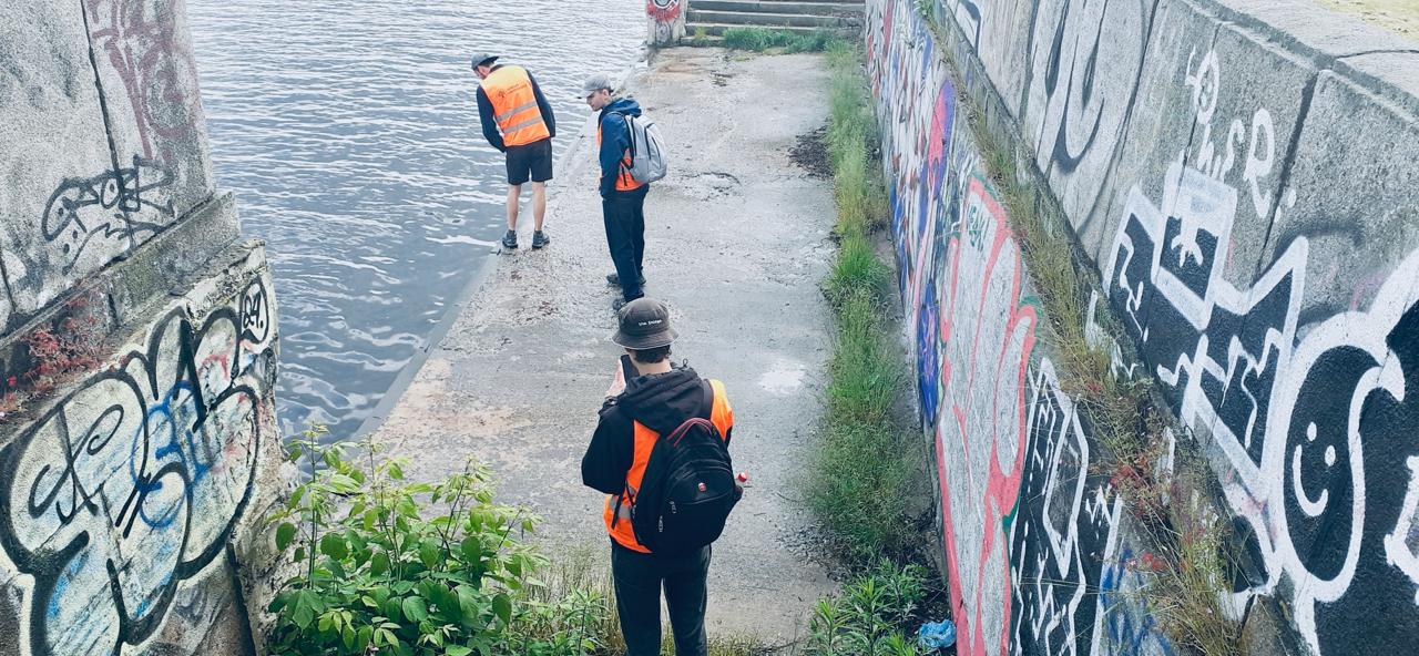 Attēlā redzami trīs studenti, kuri atrodas pie betonētas ūdenstilpnes malas. Betona sienas ir apzīmētas ar dažādu krāsu grafiti zīmējumiem.