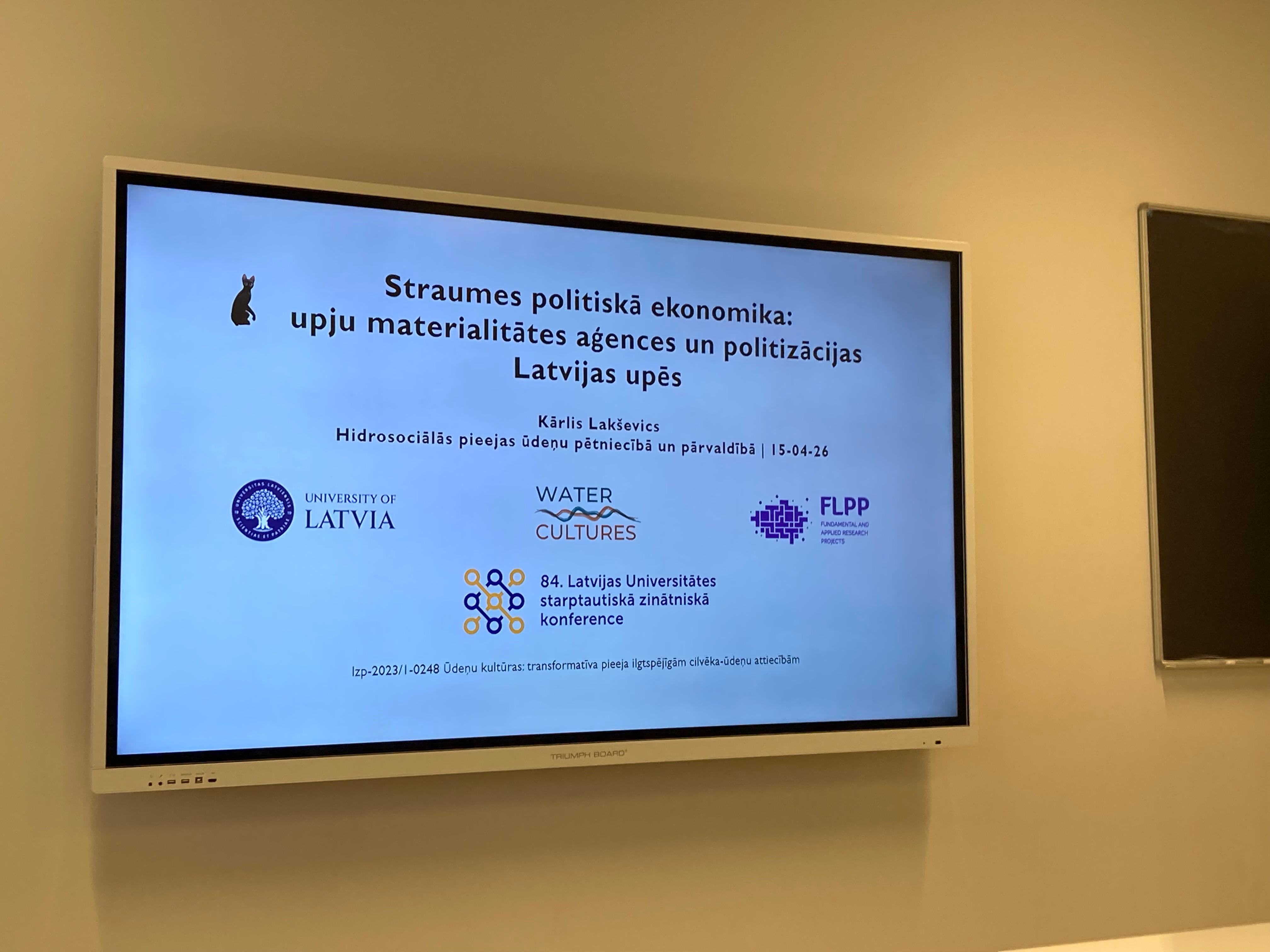 Attēlā redzams prezentācijas ekrāns ar titulslaidu. Prezentācijas nosaukums ir "Straumes politiskā ekonomika: upju materialitātes aģences un politizācijas Latvijas upēs".