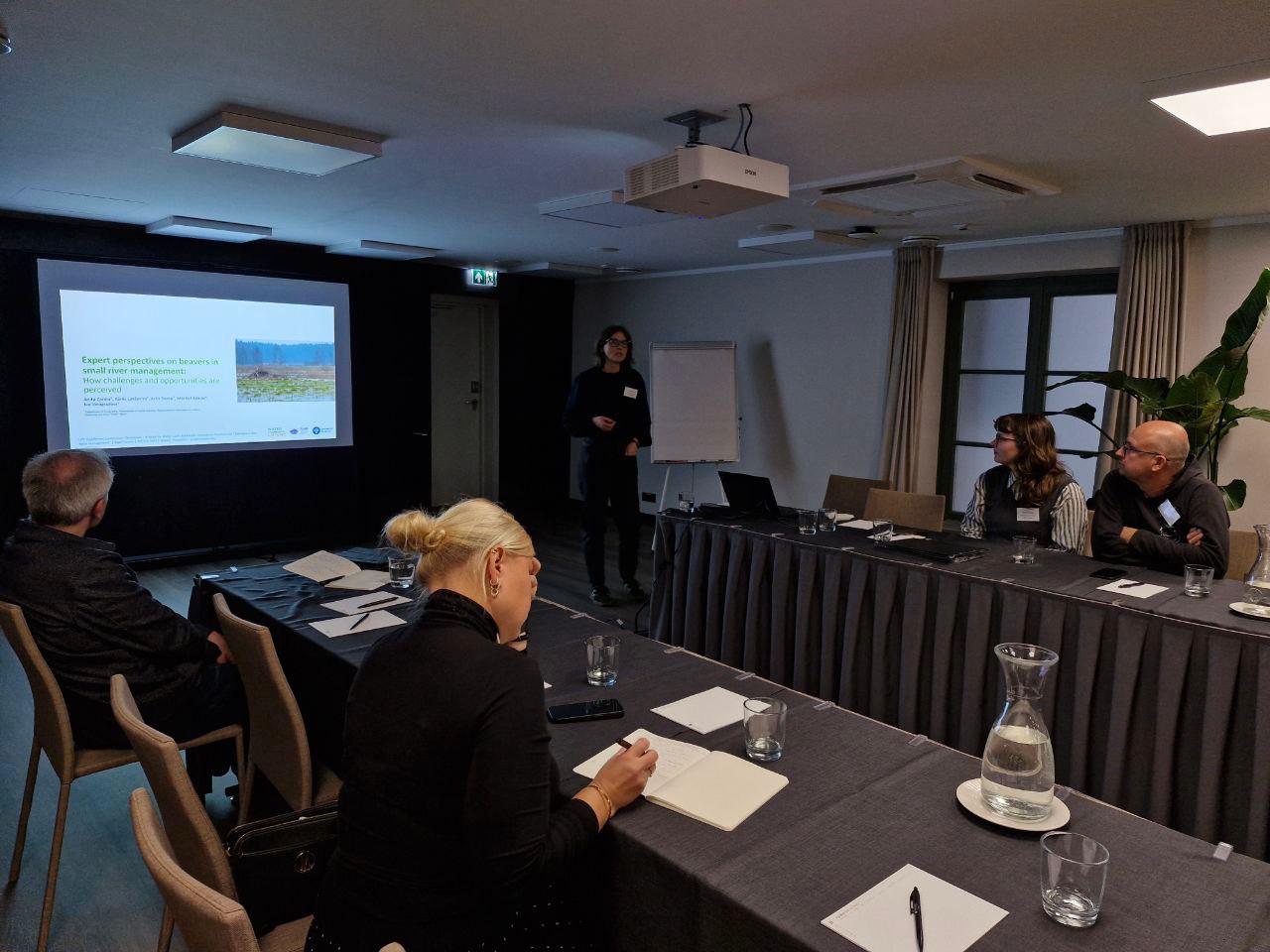 Attēlā redzama referāta prezentēšana. Kreisajā pusē redzams prezentācijas slaids ar tekstu. Priekšplānā un labajā malā galds, ap kuru sēž cilvēki. Attēla centrālajā, tālakajā daļā redzama projekta pētniece Anita Zariņa.